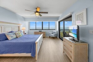 Ein Schlafzimmer mit einem Bett, einem Schminktisch, einem Sessel, einem Fernseher, einem Deckenventilator und einem großen Fenster mit Meerblick.