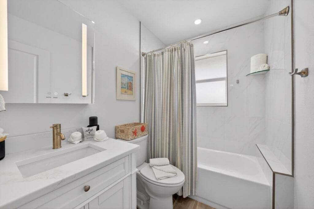 Un baño con lavamanos, inodoro y bañera blancos. Hay una cortina de ducha a rayas, un pequeño estante con una toalla y un cuadro en la pared.