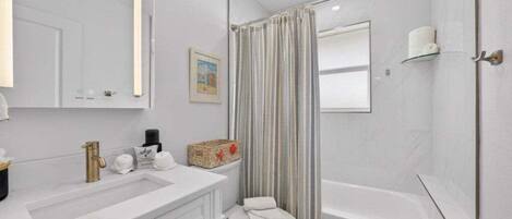 Un baño con lavamanos, inodoro y bañera blancos. Hay una cortina de ducha a rayas, un pequeño estante con una toalla y un cuadro en la pared.