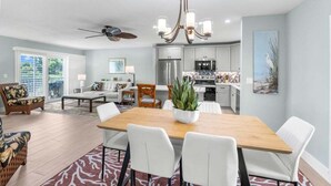 A spacious living room with a wooden dining table, white chairs, a ceiling fan, and a chandelier. - Seashells Unit 06 2 Bedroom Condo (Sanibel)