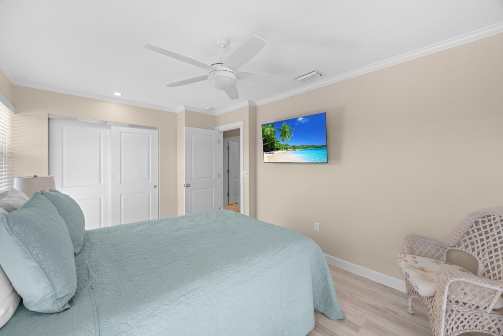 A bedroom with a bed, a ceiling fan, a TV on the wall, and a wicker chair.