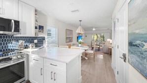 Una cocina moderna con armarios blancos, horno integrado y microondas. Cuenta con un comedor con mesa y sillas, y un cuadro grande en la pared.