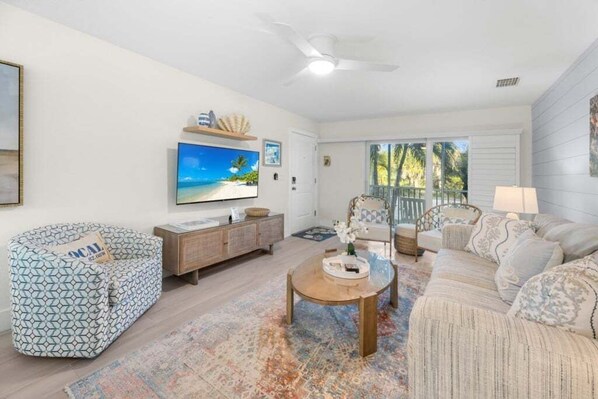 A living room with a sofa, a coffee table, a TV, and a window with a view of trees. - Seashells Unit 09 2 Bedroom Condo (Sanibel)