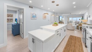 A modern kitchen with white cabinetry, a central island, pendant lighting, and a view to the living area. - Sandy Bend 1 2 Bedroom Condo (Sanibel)