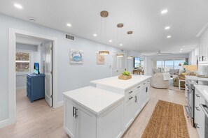 A modern kitchen with white cabinetry, a central island, pendant lighting, and a view to the living area. - Sandy Bend 1 2 Bedroom Condo (Sanibel)