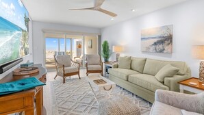 A spacious living room with a large window, a ceiling fan, a sofa, and a coffee table. - Sandy Bend 1 2 Bedroom Condo (Sanibel)