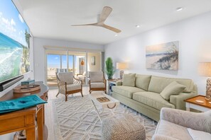 A spacious living room with a large window, a ceiling fan, a sofa, and a coffee table. - Sandy Bend 1 2 Bedroom Condo (Sanibel)