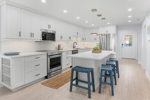 A modern kitchen with white cabinets, stainless steel appliances, and blue bar stools. - Sandy Bend 1 2 Bedroom Condo (Sanibel)