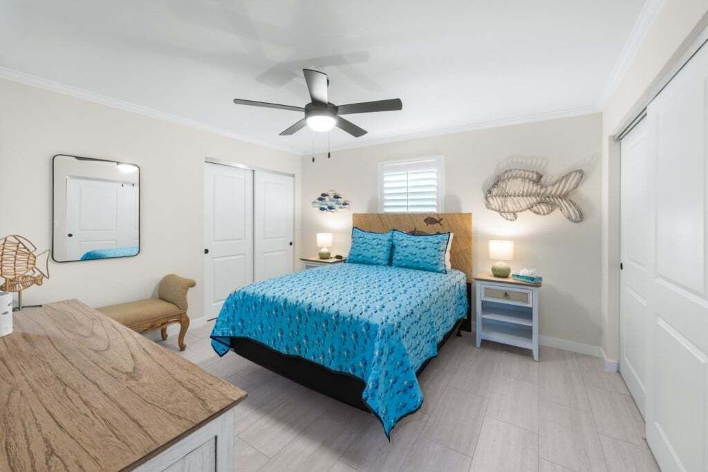 Un dormitorio con cama, ventilador de techo, mesita de noche con lámpara, una silla y una escultura de pez montada en la pared.