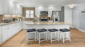 A modern kitchen with white cabinets, a central island with bar stools, a microwave, a refrigerator, and a brick accent wall. - Blind Pass Unit G201 2 Bedroom Condo (Sanibel)