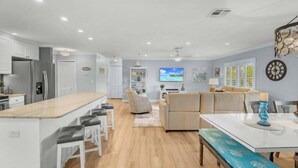 A modern kitchen with a central island, bar stools, a dining table with a bench, and a living area with a sofa and a TV. - Blind Pass Unit G201 2 Bedroom Condo (Sanibel)