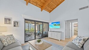A modern living room with a wooden ceiling, a large TV, and a sliding glass door leading to an outdoor area. - Coquina 3f 3 Bedroom Condo (Sanibel)