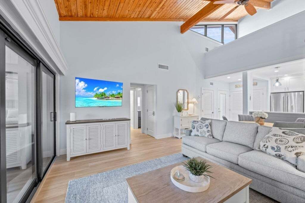 A spacious living area with a large flat-screen TV, a wooden ceiling, and a view of the ocean through large windows.