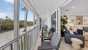 A balcony with a view of palm trees, a seating area with a sofa and a lamp, and a framed artwork on the wall. - Seashells Unit 36 2 Bedroom Condo (Sanibel)