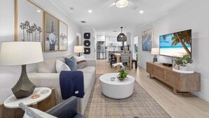 A modern living room with a sofa, coffee table, and TV. There is a kitchen in the background and a dining area. - Seashells Unit 36 2 Bedroom Condo (Sanibel)