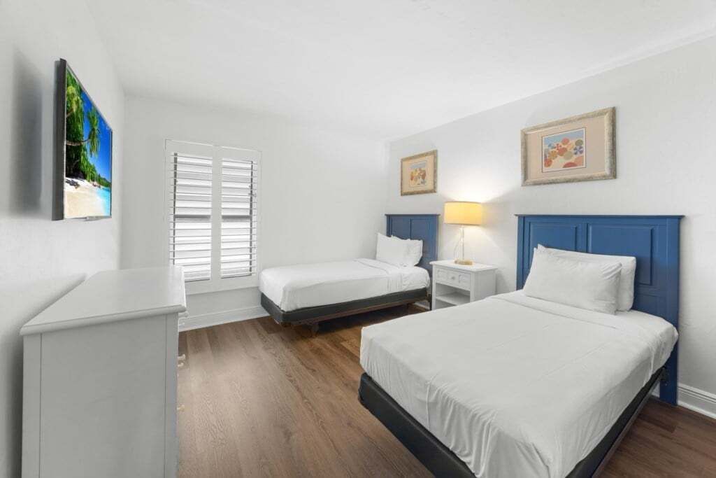 A hotel room with two single beds, a desk, a chair, a window with blinds, and two framed pictures on the wall.