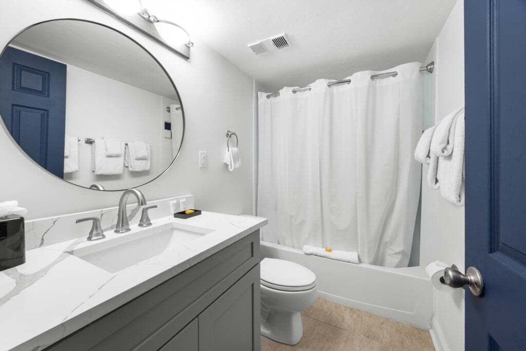 A modern bathroom with a white sink, a round mirror, a toilet, and a bathtub with a white shower curtain.