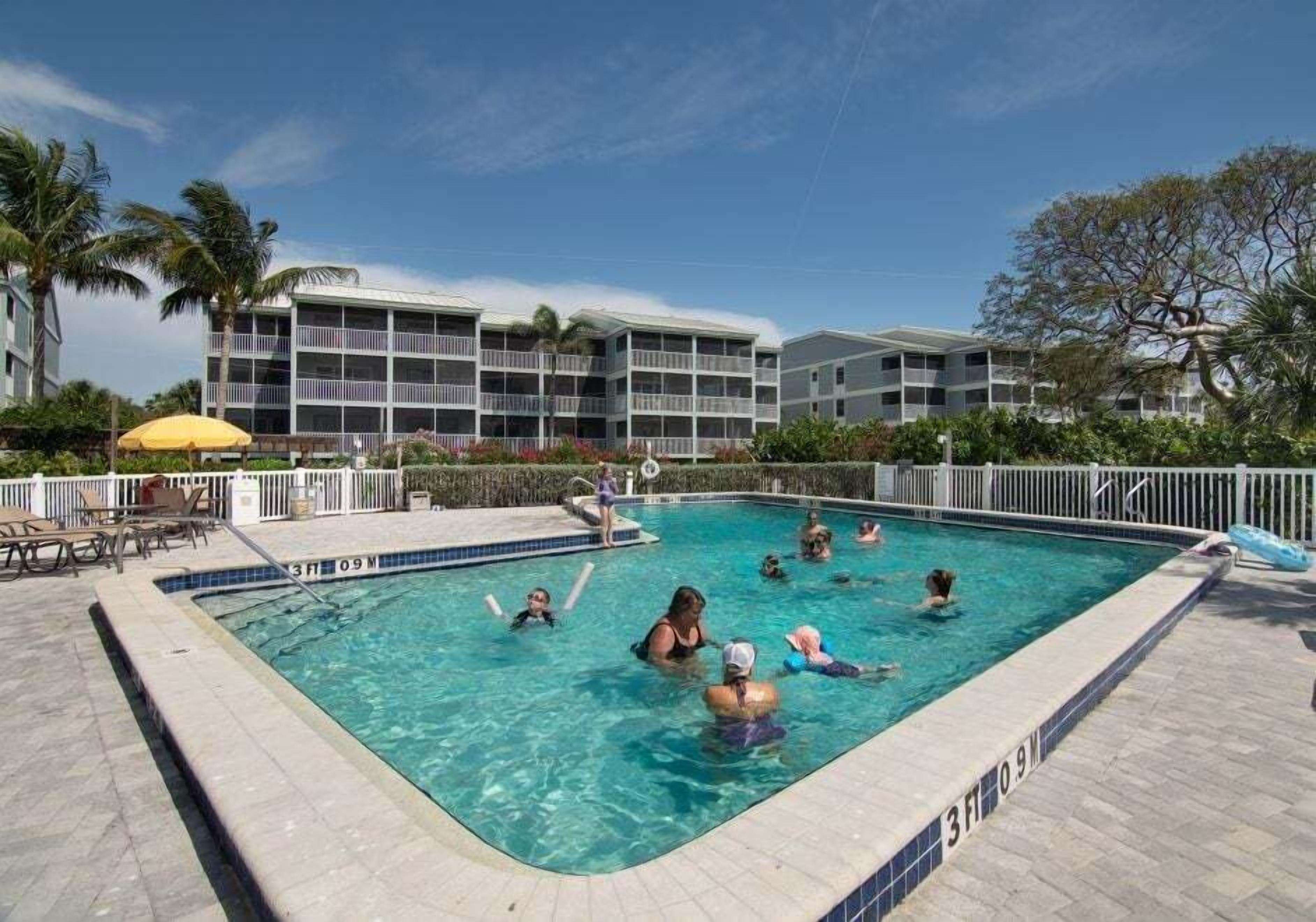 A swimming pool with people in the water, surrounded by a white fence, palm trees, and a building with balconies.