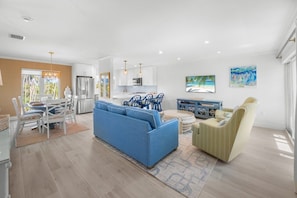 A modern living room with a blue sofa, dining area with white chairs, and a kitchen in the background. - Seashells Unit 20 - Shiny Star 2 Bedroom Condo (Sanibel)