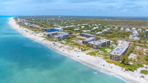 Une vue aérienne d’une zone côtière avec une plage, plusieurs bâtiments et un ciel dégagé.