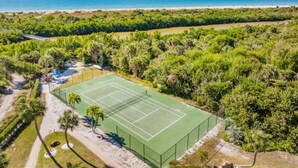 Vista aérea de una cancha de tenis rodeada de vegetación exuberante y una playa al fondo.