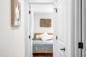 Ein schmaler Flur führt zu einem Schlafzimmer mit einem Bett, einem Wandspiegel und einer Wanddekoration.