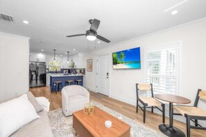 A modern living room with a sofa, coffee table, and chairs, a kitchen in the background, and a ceiling fan. - Chic Island Haven - Seashells Unit #38 2 Bedroom Condo (Sanibel)