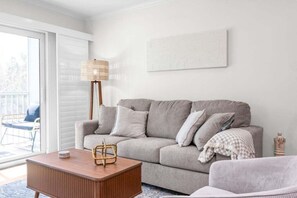 A living room with a grey sofa, a wooden coffee table, a wicker floor lamp, and a view of the outdoors through a sliding glass door. - Chic Island Haven - Seashells Unit #38 2 Bedroom Condo (Sanibel)