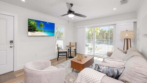 A spacious living room with a ceiling fan, a large TV on the wall, a sofa, a coffee table, and a view of a balcony with outdoor furniture. - Chic Island Haven - Seashells Unit #38 2 Bedroom Condo (Sanibel)