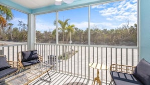 A covered outdoor area with a ceiling fan, two wicker chairs with cushions, a small table with a lamp, and large windows offering a view of the beach and palm trees. - Chic Island Haven - Seashells Unit #38 2 Bedroom Condo (Sanibel)