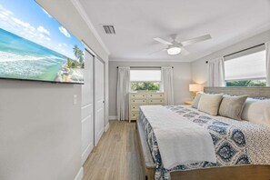 A bedroom with a bed, a nightstand, a window with curtains, a ceiling fan, and a view of the ocean. - Seashells Unit 18-coastal Breeze 2 Bedroom Condo (Sanibel)