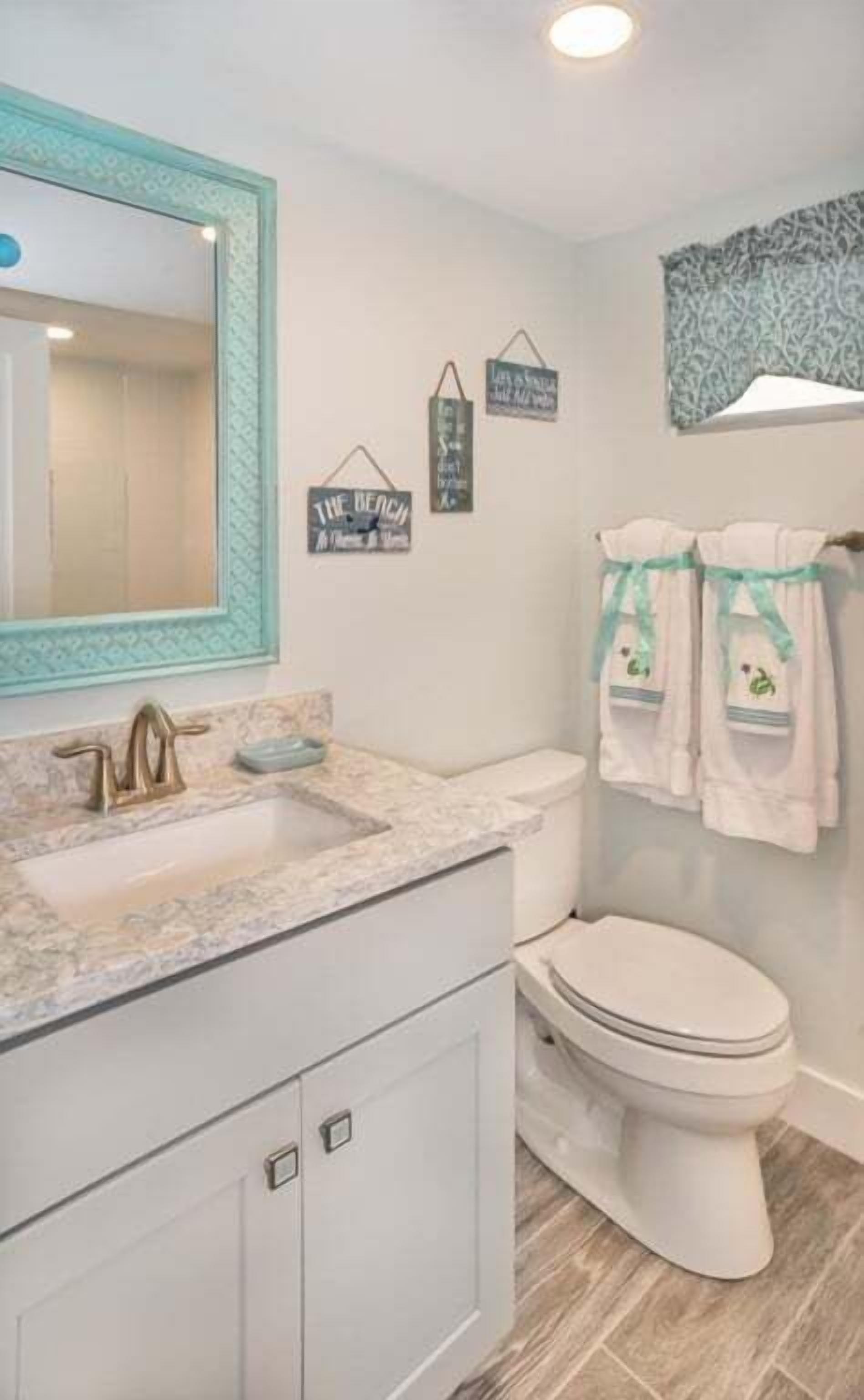 Une salle de bain avec un lavabo en marbre, des toilettes blanches et un miroir doté d’un cadre décoratif. Deux panneaux sont fixés au mur : l’un indique « The Beach » et l’autre porte un message sur l’importance de savoir lâcher prise.