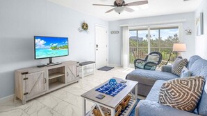 A living room with a television, a ceiling fan, a sofa, and a view of the outdoors through a sliding glass door. - Seashells Unit 44 2 Bedroom Condo (Sanibel)