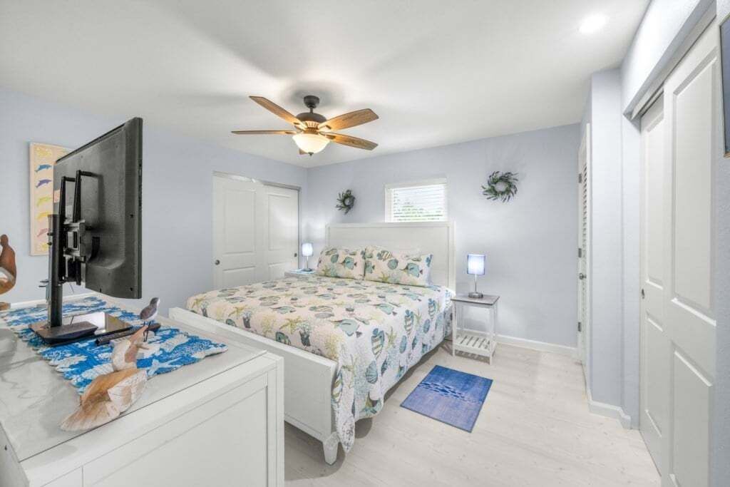 Une chambre à coucher avec un lit, un bureau avec un ordinateur, un ventilateur de plafond et une fenêtre avec des stores.