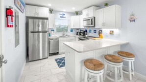 A modern kitchen with white cabinets, stainless steel appliances, a central island with stools, and a blue and white backsplash. - Seashells Unit 44 2 Bedroom Condo (Sanibel)