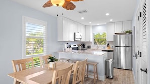 A modern kitchen with white cabinets, a stainless steel refrigerator, and a wooden dining table with chairs. - Seashells Unit 12 2 Bedroom Condo (Sanibel)