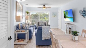 A modern living room with a blue sofa set, a dining table, and a TV on the wall. - Seashells Unit 12 2 Bedroom Condo (Sanibel)