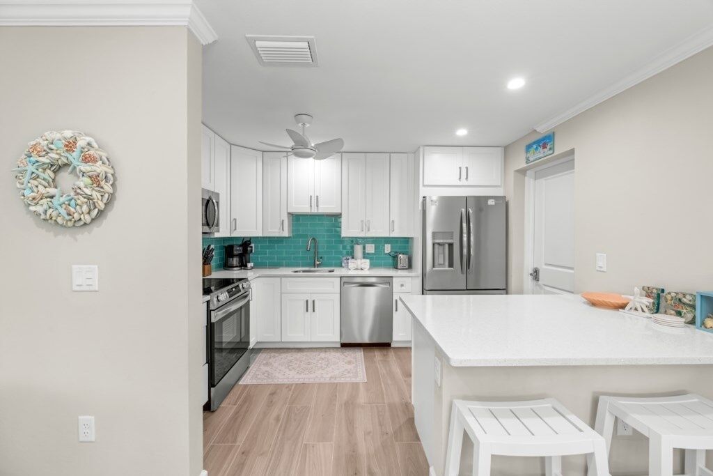 A modern kitchen with white cabinets, a teal backsplash, stainless steel appliances, and a white island with stools.