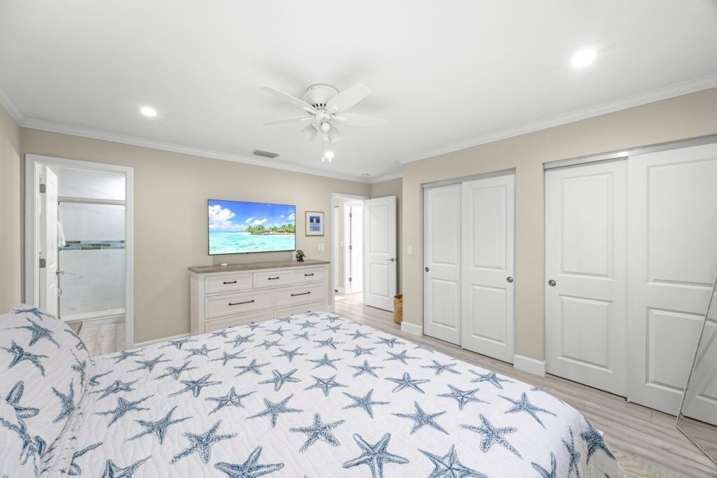 A bedroom with a bed, a television, a ceiling fan, and a view of the beach through the window.