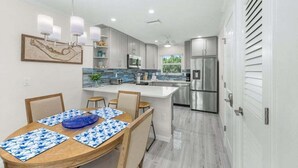 A modern kitchen with a central island, stainless steel appliances, and a dining area with a wooden table and blue placemats. - Seashells Unit 33 2 Bedroom Condo (Sanibel)