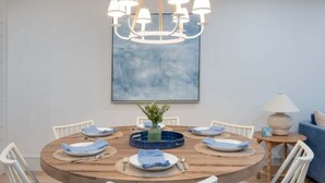 A round wooden dining table with white chairs, a chandelier, and a blue abstract painting above. - Sanddollar A302 3 Bedroom Condo (Sanibel)