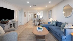 A modern living room with a sofa, coffee table, and TV. There is a dining area with chairs and a table. - Sanddollar A302 3 Bedroom Condo (Sanibel)