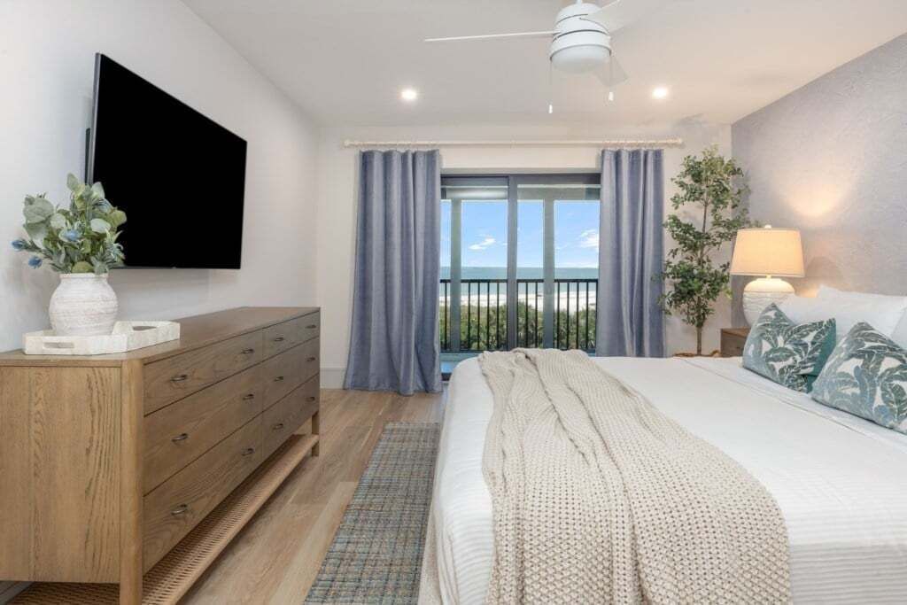 Et soveværelse med seng, kommode, fjernsyn ophængt på væggen, en balkon med gardiner og udsigt til det fri.
