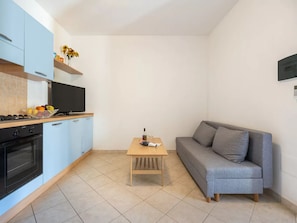 A compact kitchen with a stove, oven, and blue cabinets. A small wooden table with a bottle and glasses is in the center. A gray sofa is against the wall. - Comfortable Accommodation With Patio (Badesi)