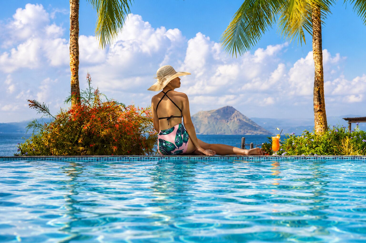 A person sitting by a pool with a view of the ocean and a mountain in the background.