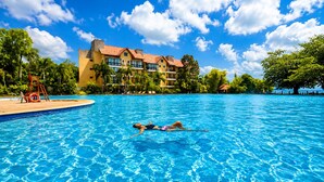 A swimming pool with two people swimming, surrounded by a hotel building with multiple balconies and palm trees.