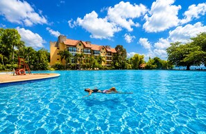 En swimmingpool med två personer som simmar, omgiven av en hotellbyggnad med flera balkonger och palmer.