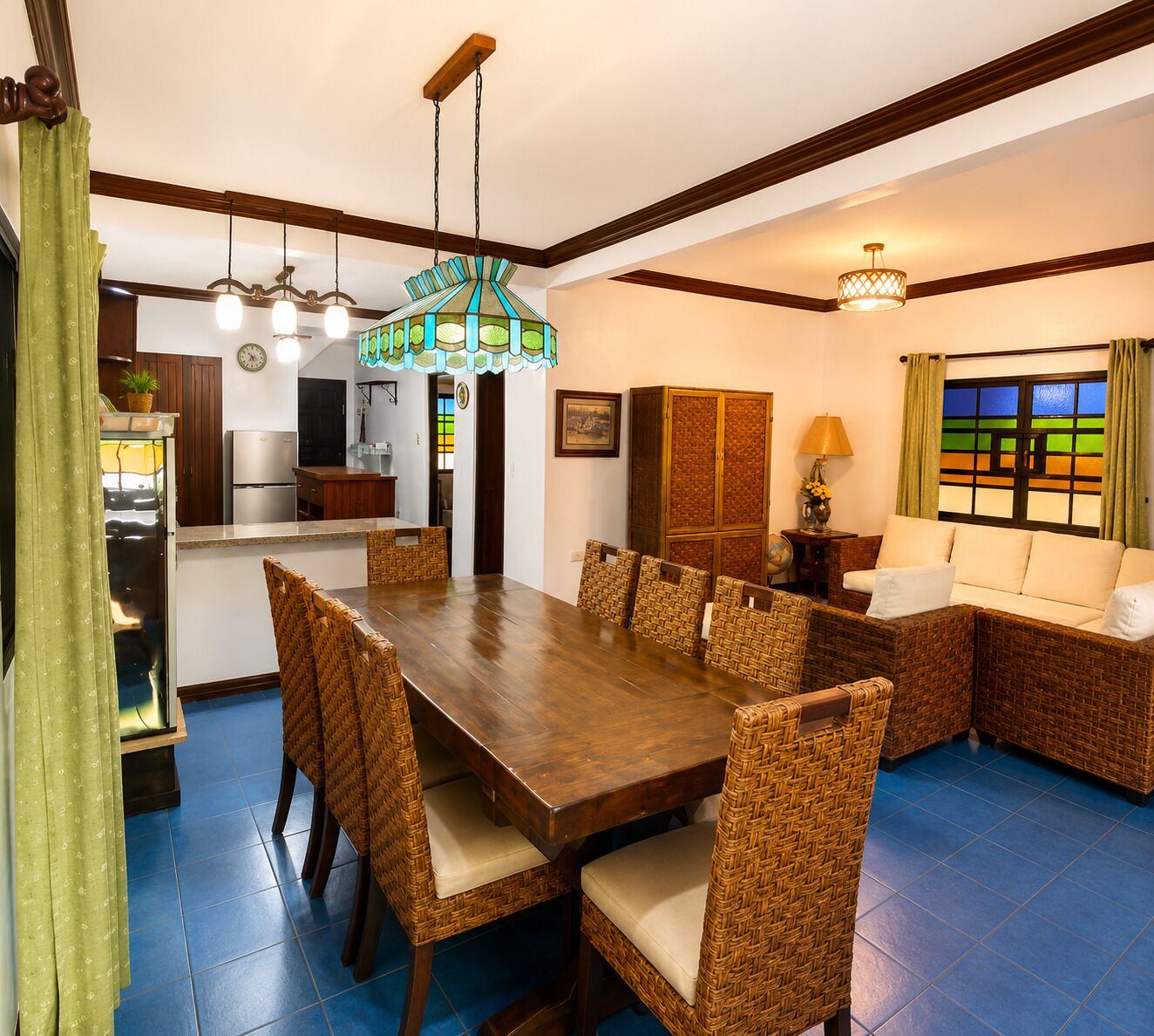 A dining area with a wooden table and wicker chairs, a kitchen in the background, and a stained glass window.