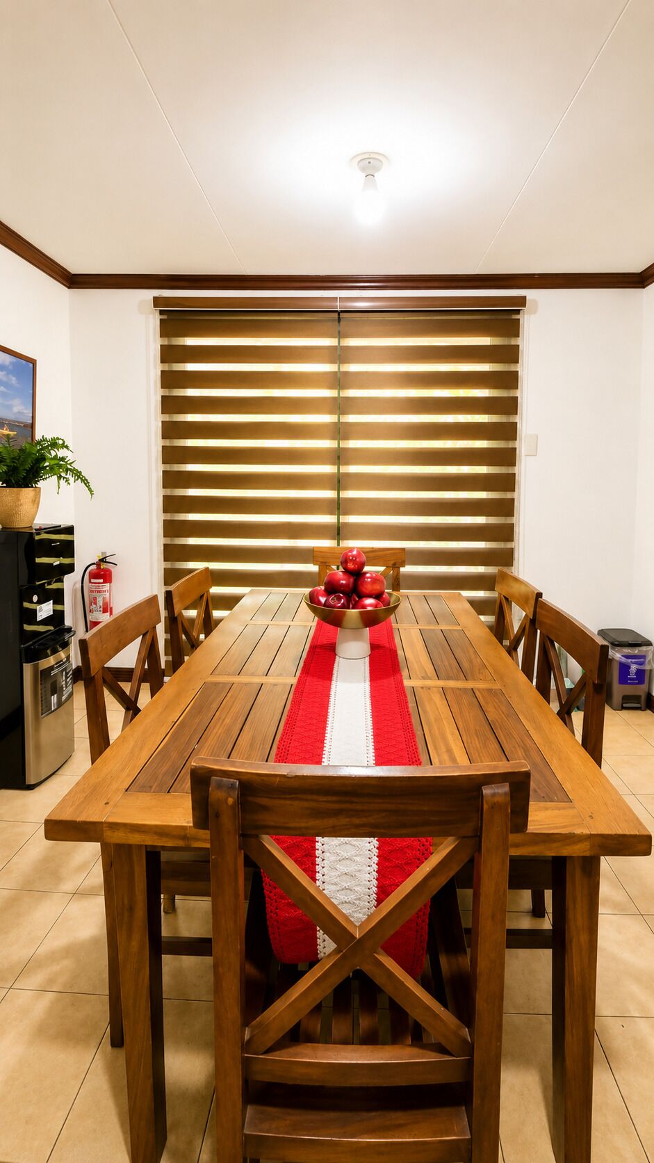 Un comedor con una mesa de madera, sillas, camino de mesa rojo y blanco, y un cuenco de manzanas.