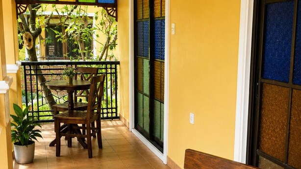 A balcony with a wooden table and chairs, a potted plant, and a view of greenery through a window with colorful panes.